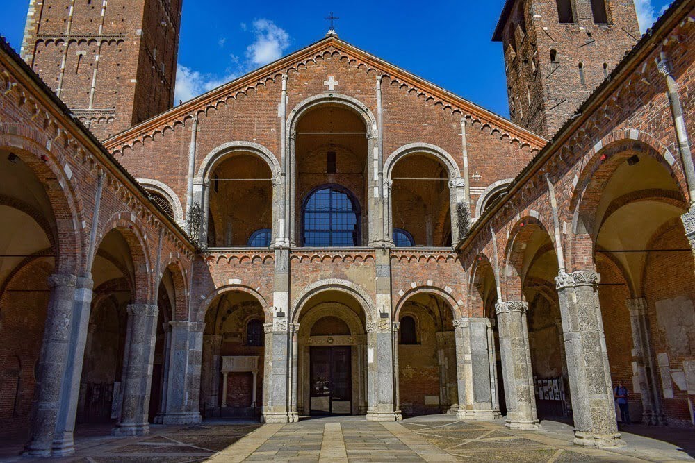 Visão frontal da Basílica de Sant'Ambrogio com vários arcos, duas torres e teto inclinado em formato de triangulo.