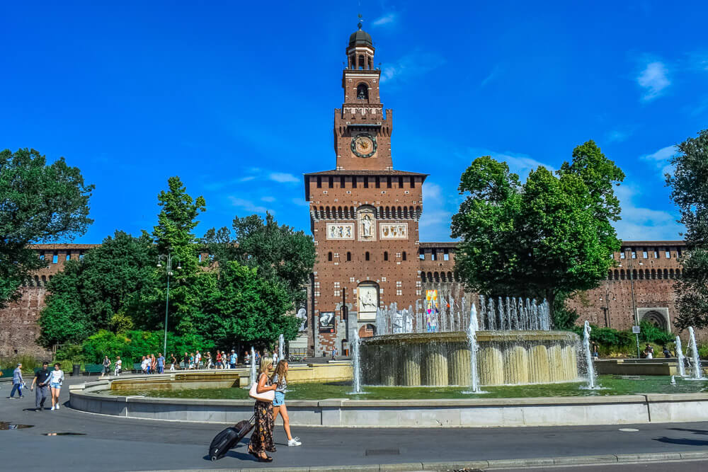 Fachada do Castelo Sforzesco com árvores e fontes em frente.
