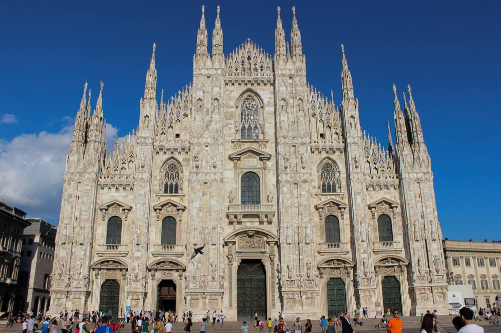 Vista frontal para a Catedral de Milão. Há um prédio religioso em tom de areia repleto de janelas, portas e fachada pontuda.