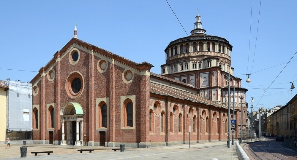 Fachada da Igreja de Santa Maria delle Grazie com paredes de tijolos avermelhados e torre majestosa ao fundo.