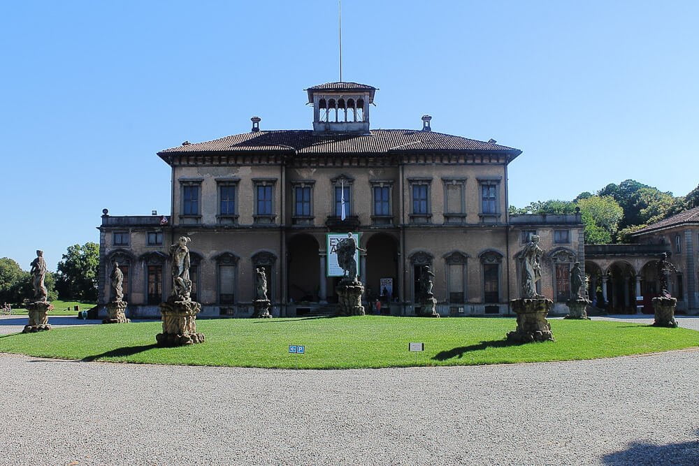 Entrada do Museu Bagatti Valsecchi com teto inclinado e esculturas no gramado da frente.