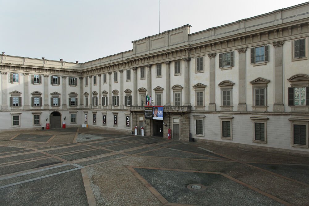 Vista parcialmente lateral do Palácio Real. Há um pátio grande em frente ao palácio.