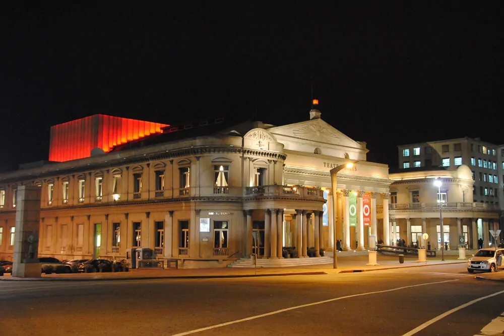 Teatro Solís à noite. Há uma rua larga e um prédio ao lado do teatro.