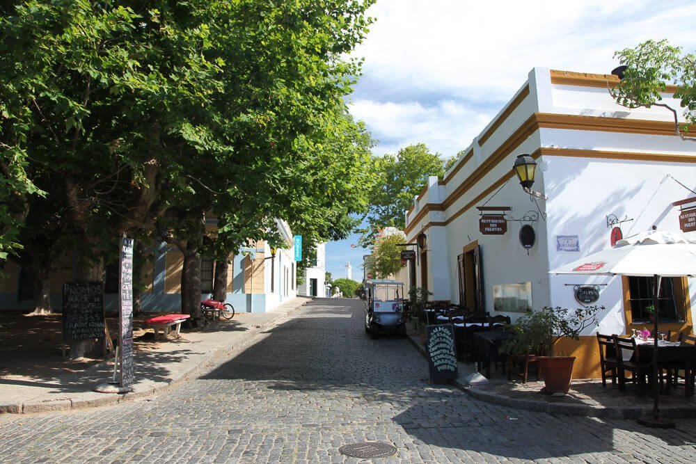 Rua com arquitetura colonial na Colônia do Sacramento. Há árvores e restaurantes agradáveis.