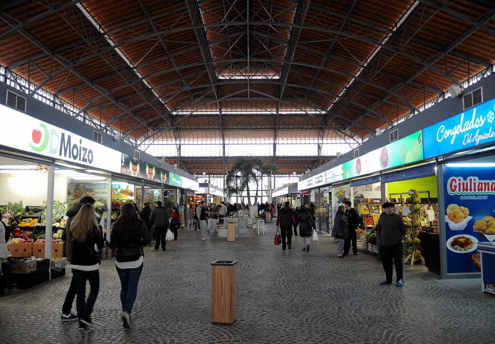 Interior do Mercado Agrícola, em Montevidéu. Há barracas com alimentos nas duas laterais do mercado.