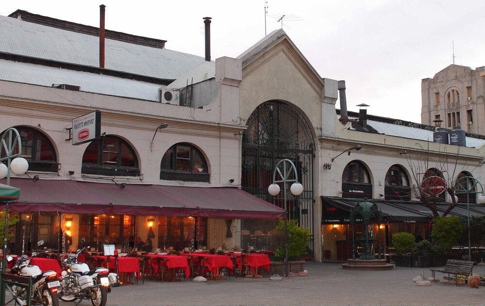 Fachada do Mercado Del Puerto. Há mesas cobertas por toalhas de mesa vermelhas.