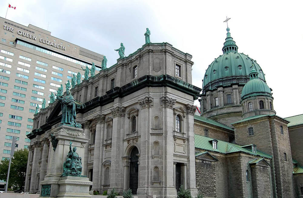 Visão externa da Cathédrale Marie-Reine-du-Monde. À esquerda, há o prédio do hotel Fairmont The Queen Elizabeth.