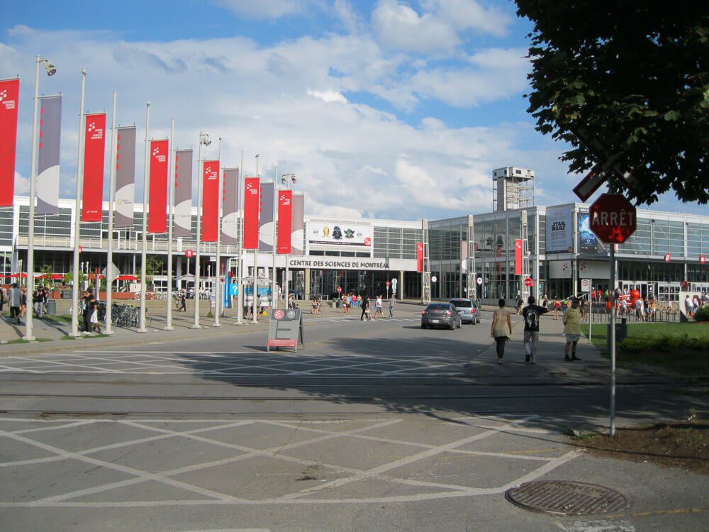 Pátio de entrada para o The Montreal Science Center. Há pessoas e carros circulando pela entrada do local.