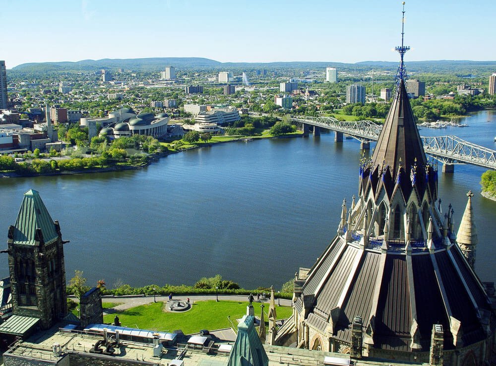 Cidade de Ottawa vista de cima. Há uma ponte sobre águas azuis, muitos prédios e áreas verdes.