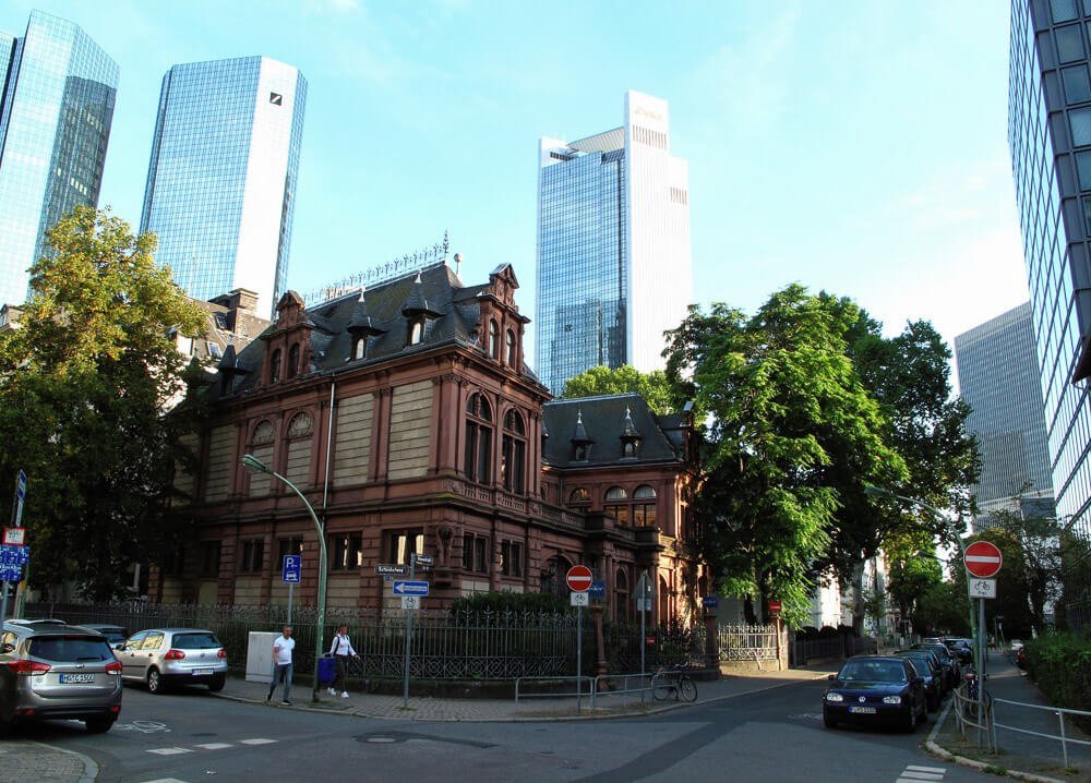 Cruzamento em Westend com casarão marrom ao centro, árvores e prédios de luxo ao fundo.