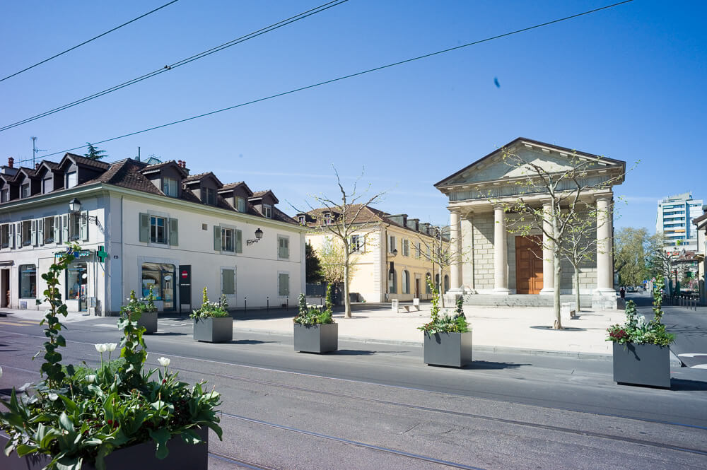Rua na região de Carouge, em Genebra. Há edifícios baixos com teto escuro e inclinado. Há também um imóvel com fachada neoclássica e paredes claras.