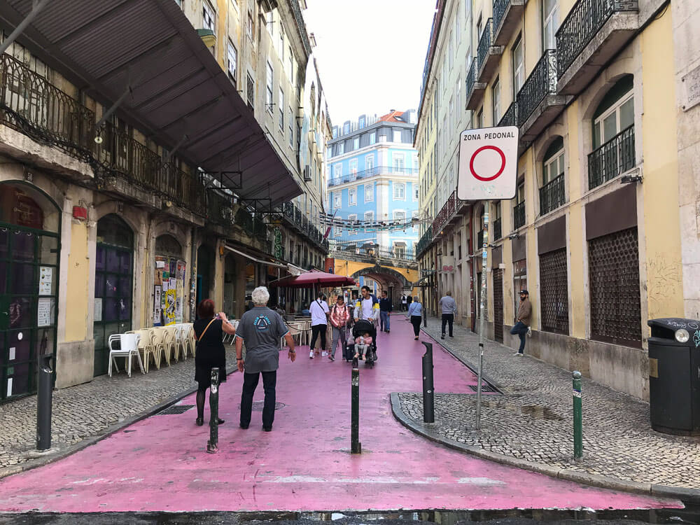 Rua Nova do Carvalho, no bairro Cais do Sodré. Há bares à esquerda e uma ponte amarela no fim da rua.