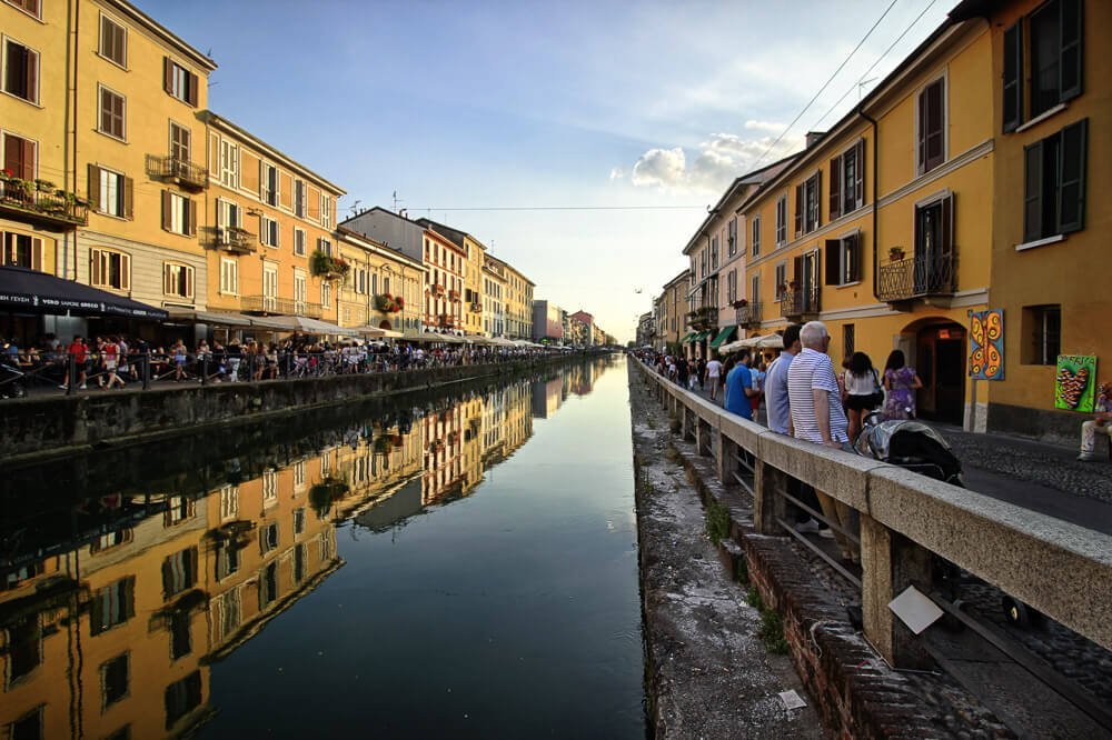 Águas do Canal Navigli refletindo o azul do céu. Há pessoas nos restaurantes às margens do canal.