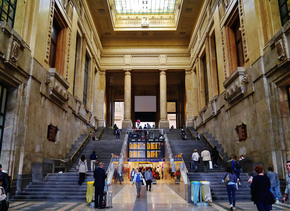 Estrutura interna da Estação Central de Milão com enormes escadas laterais e colunas cilíndricas.