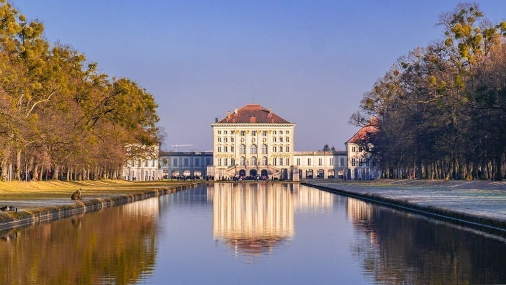 Palácio Nymphenburg com lago em frente e árvores nas laterais.