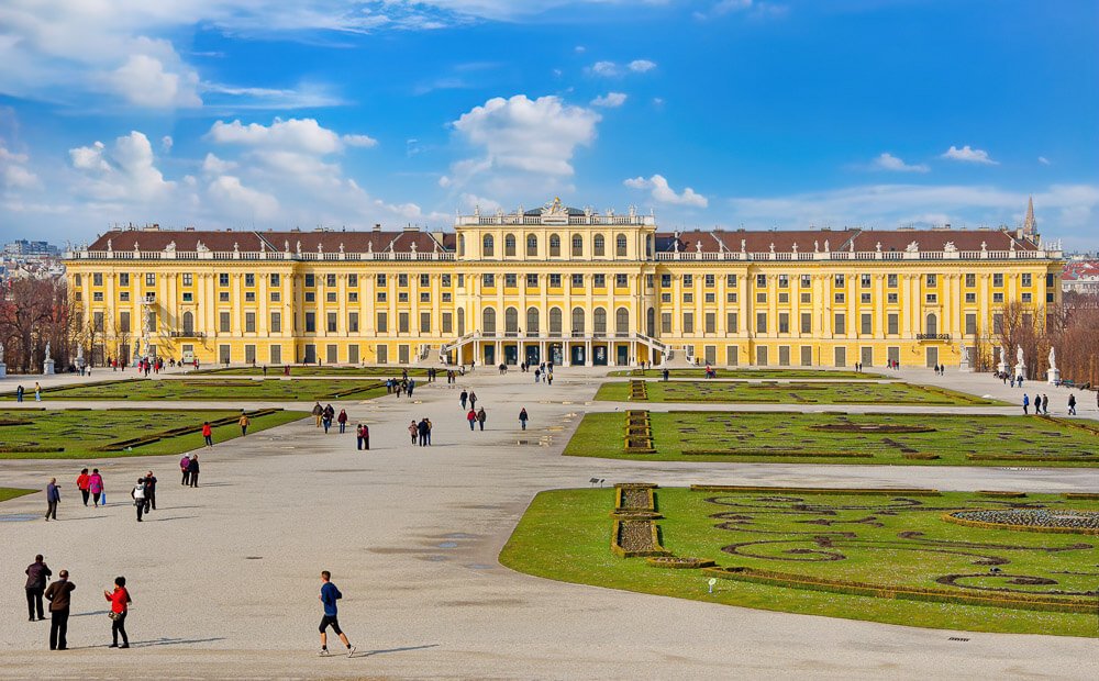 Palácio de Schönbrunn com jardins, fachada imponente e pessoas caminhando na área em frente ao local.