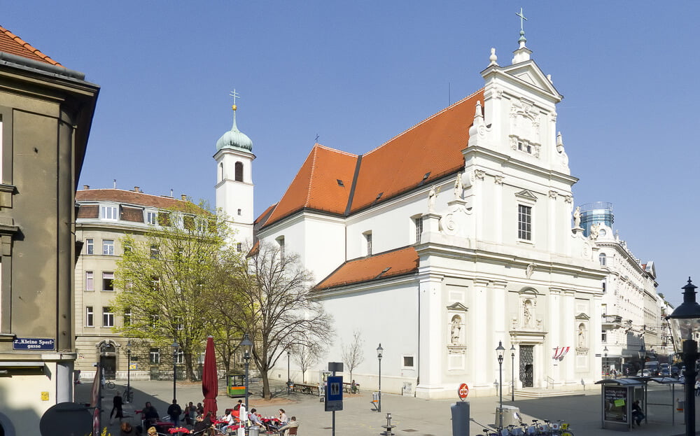 Igreja Katholische Kirche St. Josef, em Leopoldstadt. Há algumas árvores e prédios com cores claras nos arredores da igreja.