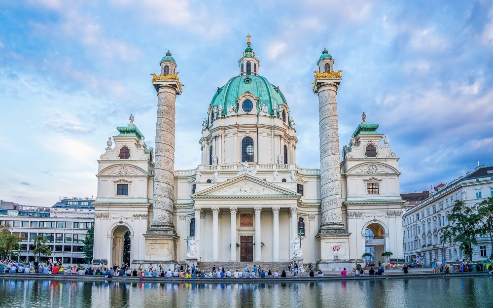 Igreja Karlskirche, em Wieden, com fachada branca com colunas cilíndricas e cúpula turquesa. Há um lago em frente à igreja.