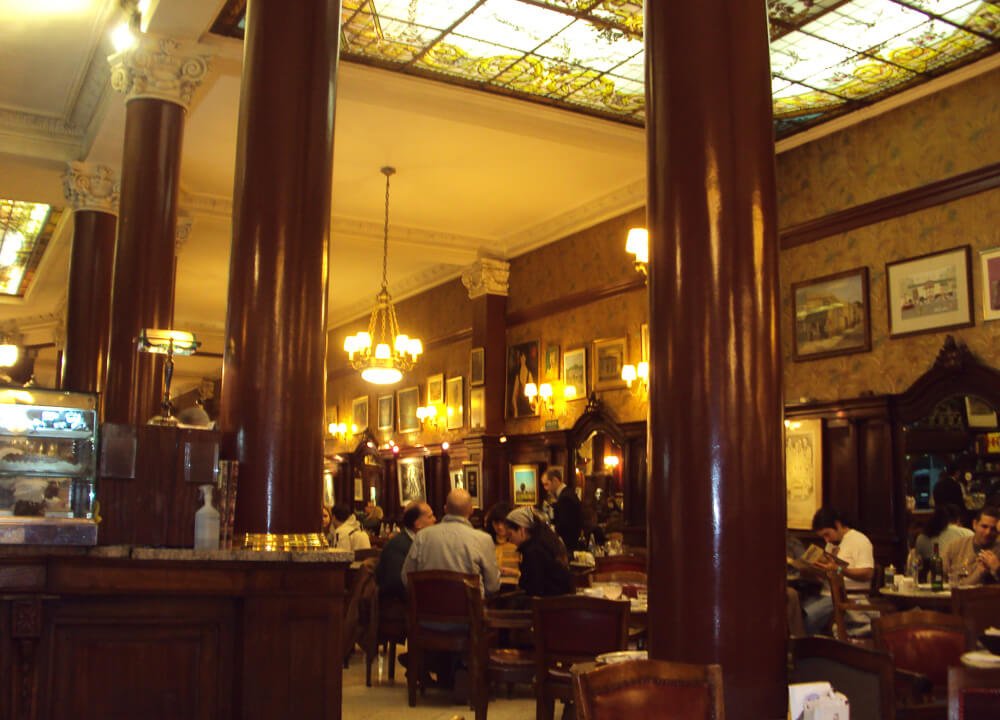 Salão interno do Café Tortoni com várias mesas, cadeiras, lustres, colunas e estufas.