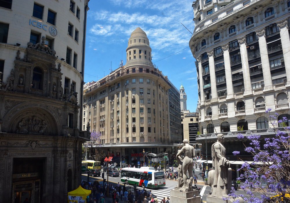 Rua Calle Florida com vários pedestres, estátuas, ônibus e edifícios nos arredores da rua.