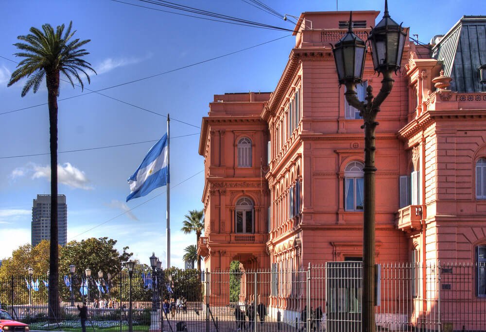 Vista lateral para o prédio da Casa Rosada, com grades ao redor do prédio e uma bandeira da Argentina hasteada.