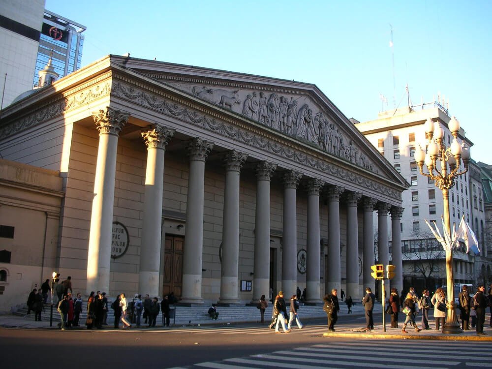 Fachada da Catedral Metropolitana de Buenos Aires com uma rua em frente e vários pedestres caminhando pelo local.