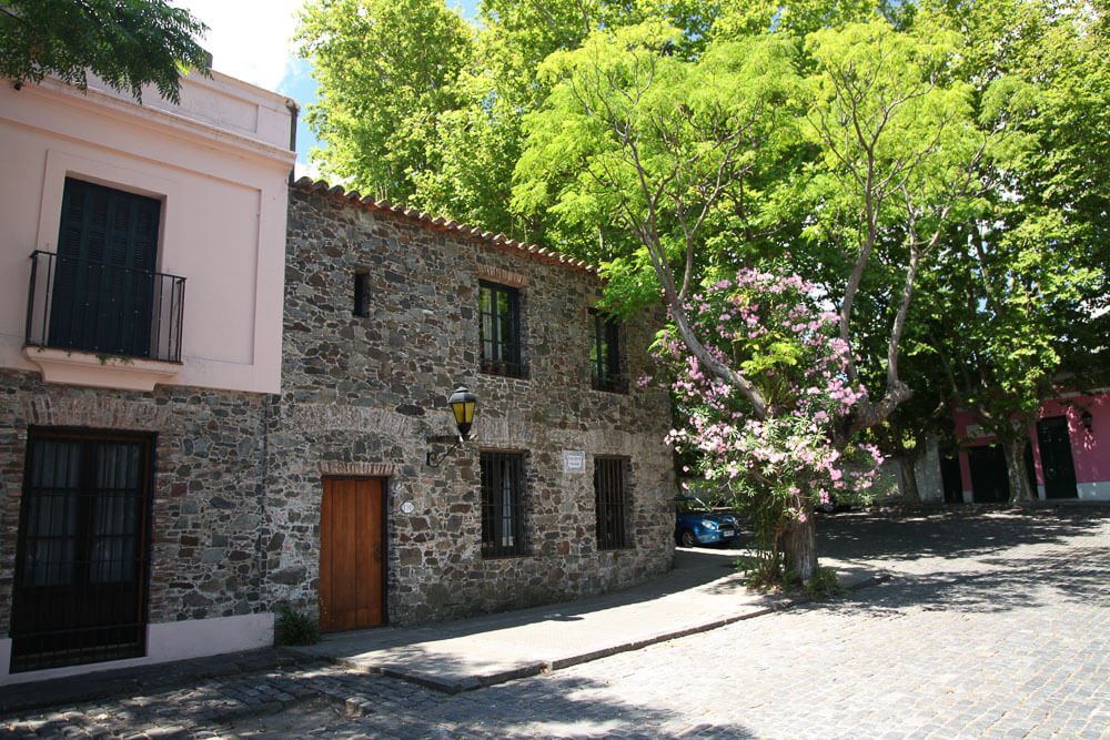 Rua na Côlonia del Sacramento, com sobrado com fachada de pedras, árvores e chão de paralelepípedos.