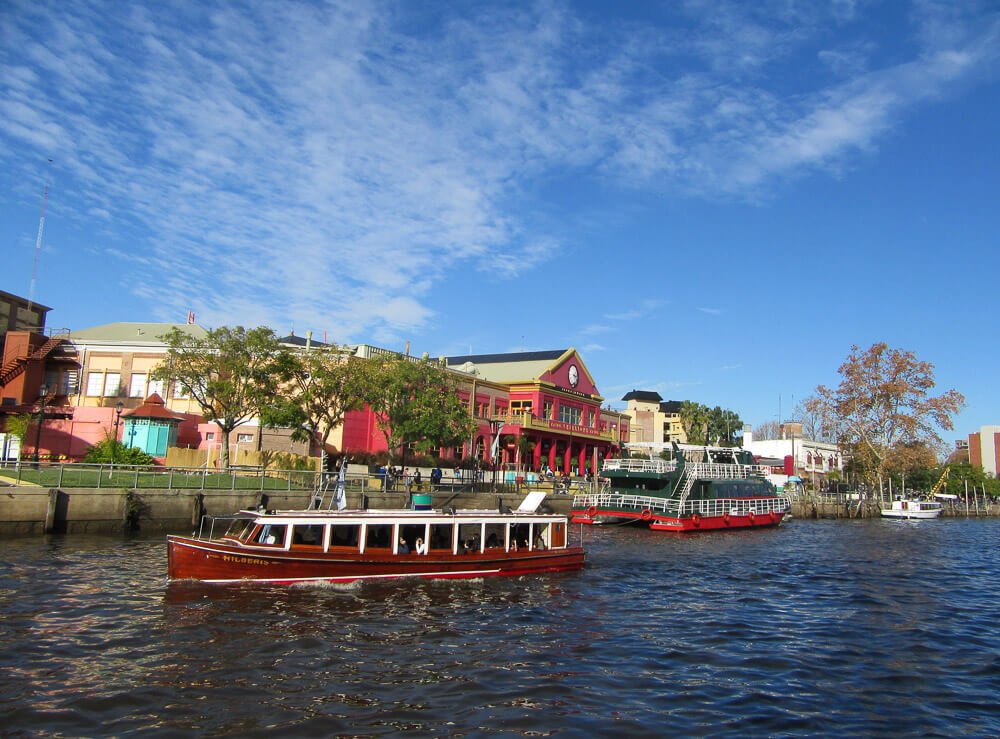 Barcos sobre o rio Delta Tigre e, ao fundo, árvores e casas na região costeira.