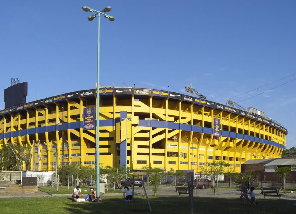 Estádio La Bombonera visto por fora, com um gramado nos arredores e pessoas caminhando próximas ao local.