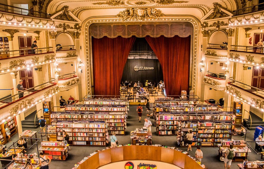 Interior da Livraria El Ateneo com muitas estantes, livros, um palco com cortina vermelha e arquibancadas elegantes nas laterais.