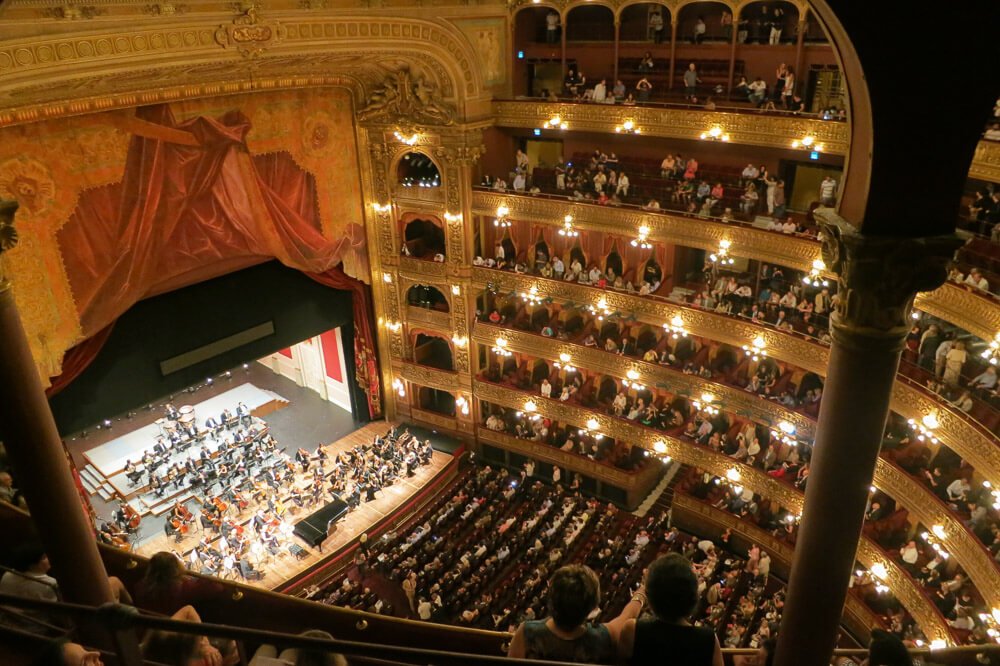 Interior do Teatro Colón com várias pessoas assistindo a uma apresentação de orquestra.