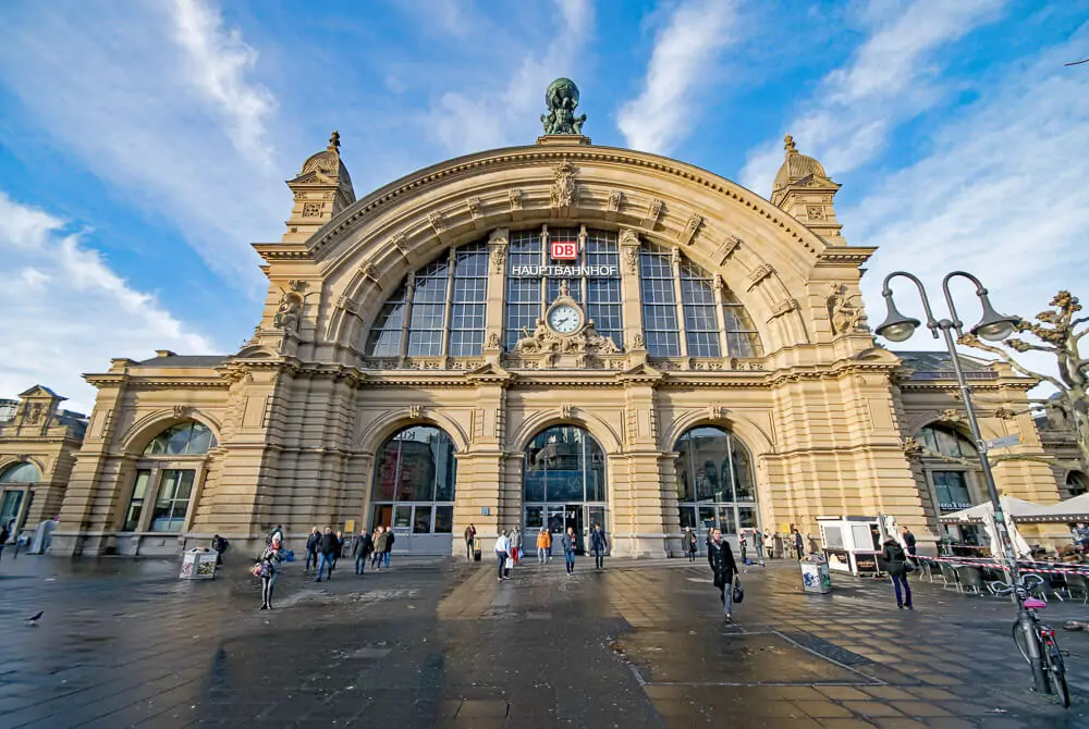 Entrada da Estação Central de Frankfurt. Há um prédio com fachada em tom de areia e pessoas transitando pelo local.