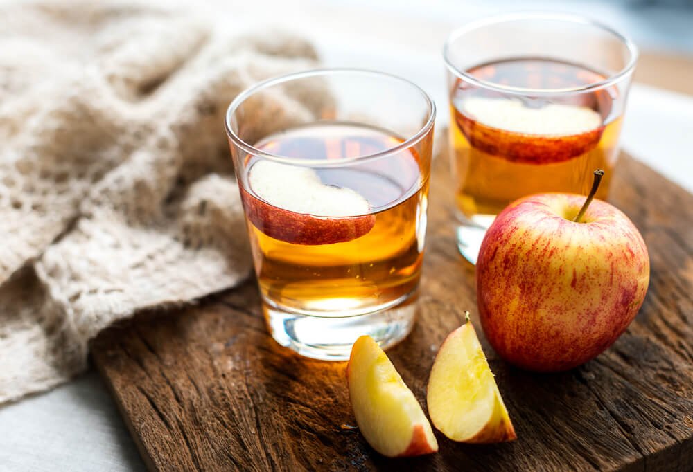 Tábua de madeira com dois copos de sidra de maçã e algumas maçãs ao lado.