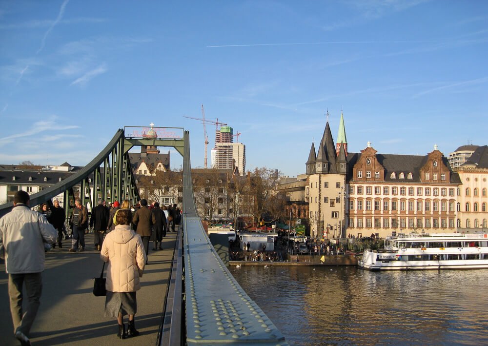 Eiserner Steg com várias pessoas caminhando sobre ela e, à direita, o Rio Meno e os edifícios de Frankfurt.