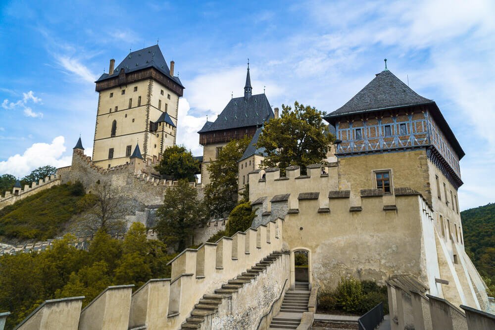 Castelo de Karlštejn com três torres, várias escadarias, paredes beges e algumas árvores.