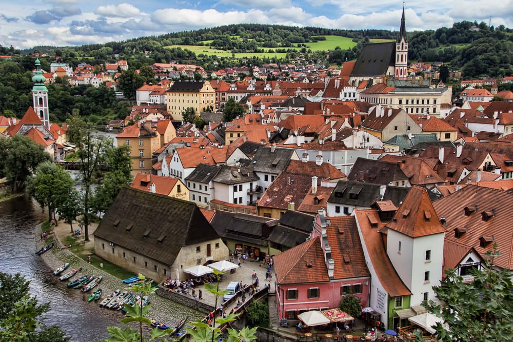 Visão superior da cidade de Český Krumlov, com construções medievais, um canal e muito verde.