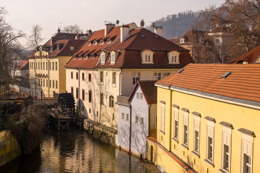 Canal Čertovka com ponte, moinho, árvores e edifícios na lateral direita.