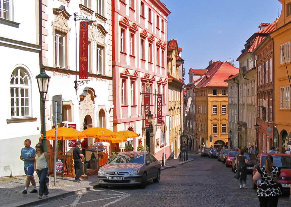 Rua com carros, chão de paralelepípedos, pessoas caminhando e edifícios graciosos e, na região de Malá Strana.