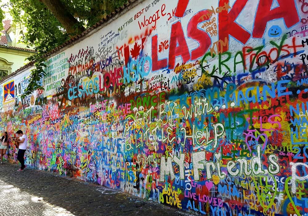 Muro de John Lennon com muitas palavras coloridas e algumas pessoas em frente ao muro.