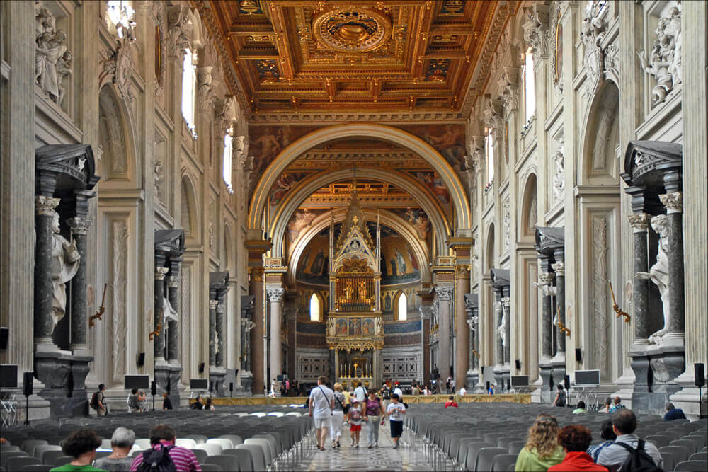 Interior da Arquibasílica de São João de Latrão com teto luxuoso, muitas estátuas nas paredes, cadeiras e pessoas.