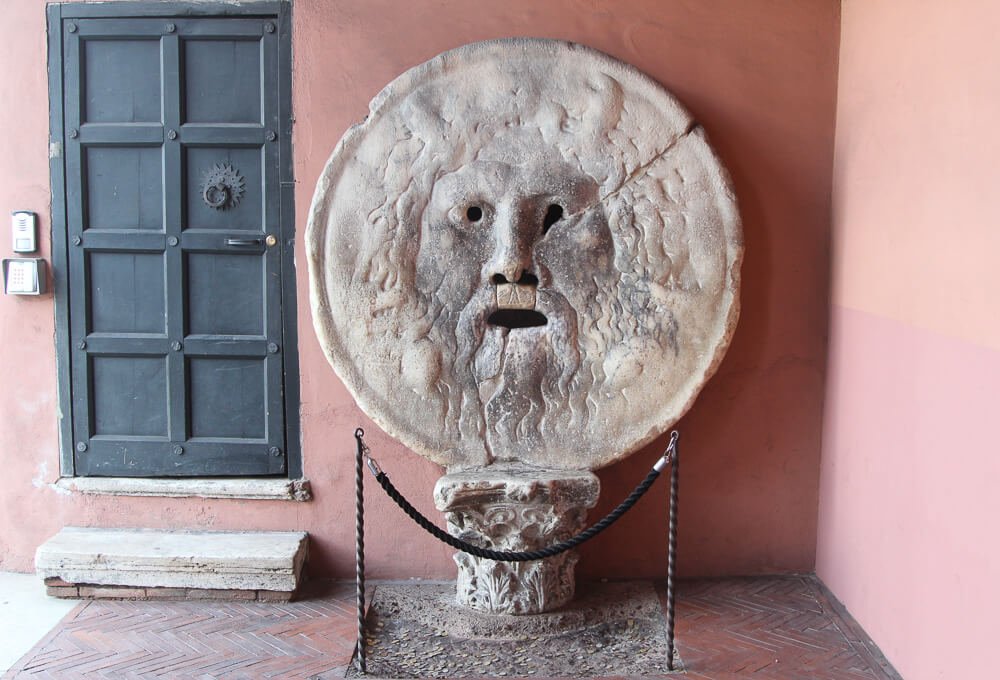 Máscara Bocca della Veritá e, ao fundo, uma parede e uma porta.