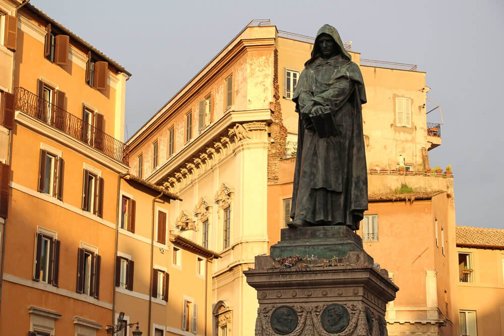 Estátua de Giordano Bruno com prédios alaranjados ao fundo.