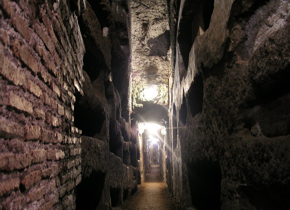 Interior das Catacumbas de Roma com paredes de pedras e corredor estreito.