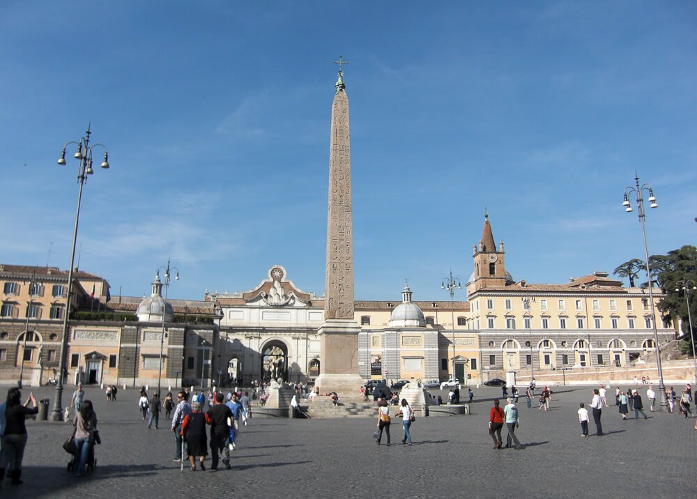 Piazza del Popolo com monumento no centro, edifícios com fachadas claras ao redor e um extenso pátio com várias pessoas caminhando.