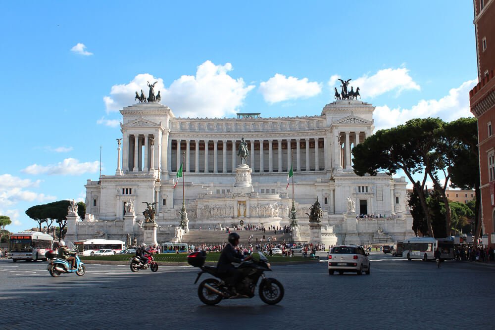 Piaza Veneza com Monumento a Vittorio Emanuele II, veículos e pessoas circulando pela praça.