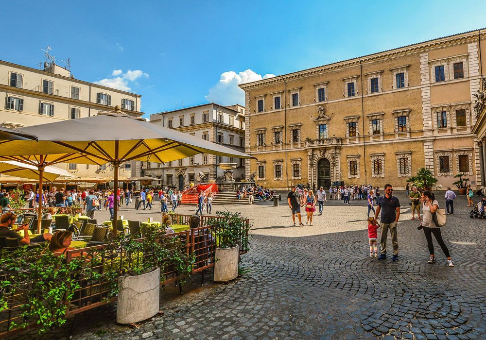 Piazza di Santa Maria in Trastevere com prédios retangulares, restaurantes e muitas pessoas.