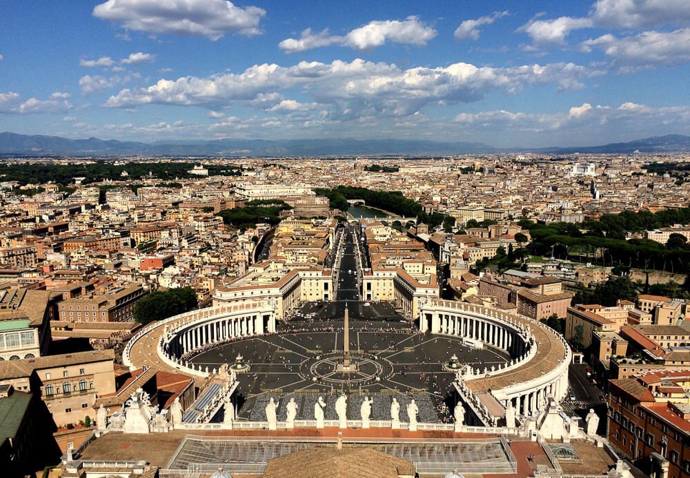 Região do Vaticano visto de cima, com a Praça de São Pedro, muitas árvores e edifícios com tetos amarronzados.