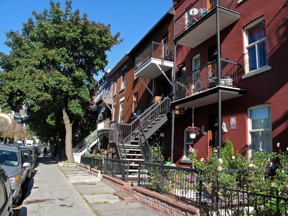 Rua com árvores e casas com jardins e escadas externas, em Mile End.