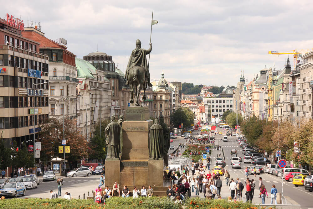 Praça Venceslau com estátua equestre no centro, pedestres no local, duas vias e prédios nas laterais.