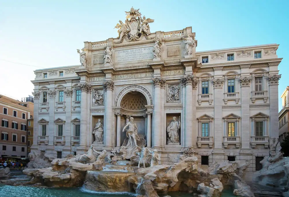 Fontana di Trevi com prédio em tom de areia, várias estátuas, pedras grandes e água azul-turquesa.